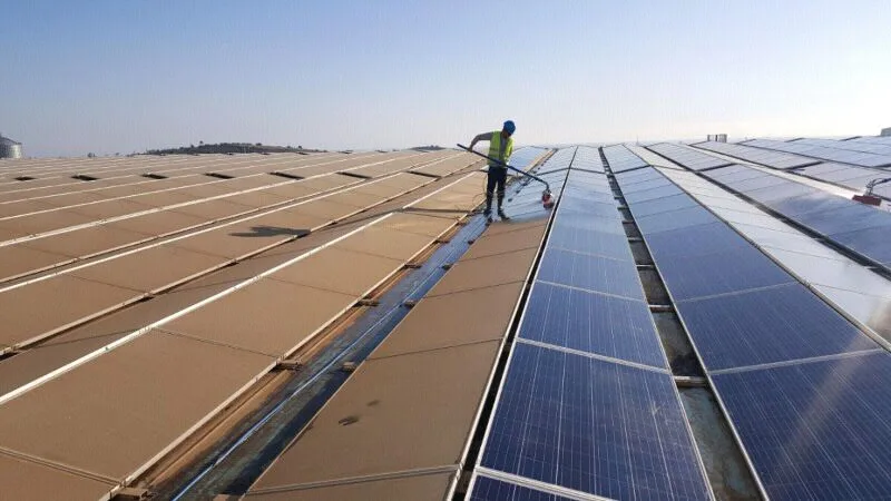 Rooftop solar PV installation mid-cleaning showing dramatic contrast between heavily dust-soiled brownish uncleaned modules on the left and freshly cleaned dark-blue modules on the right, with a worker cleaning in the middle