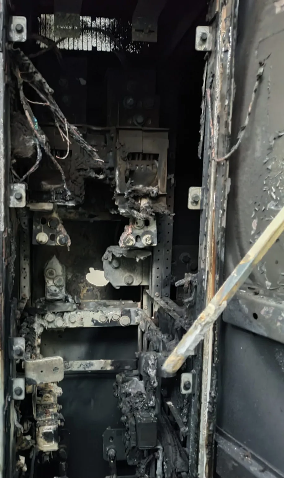 Close-up of IGBT power module section inside a fire-damaged 1 MW central solar
inverter, showing two vertical columns of metal mounting plates that clamp power
modules against the heatsink contact surface, all surfaces burned black, ash and
burned component material on the cabinet floor below, heatsink structures present
but fire-damaged