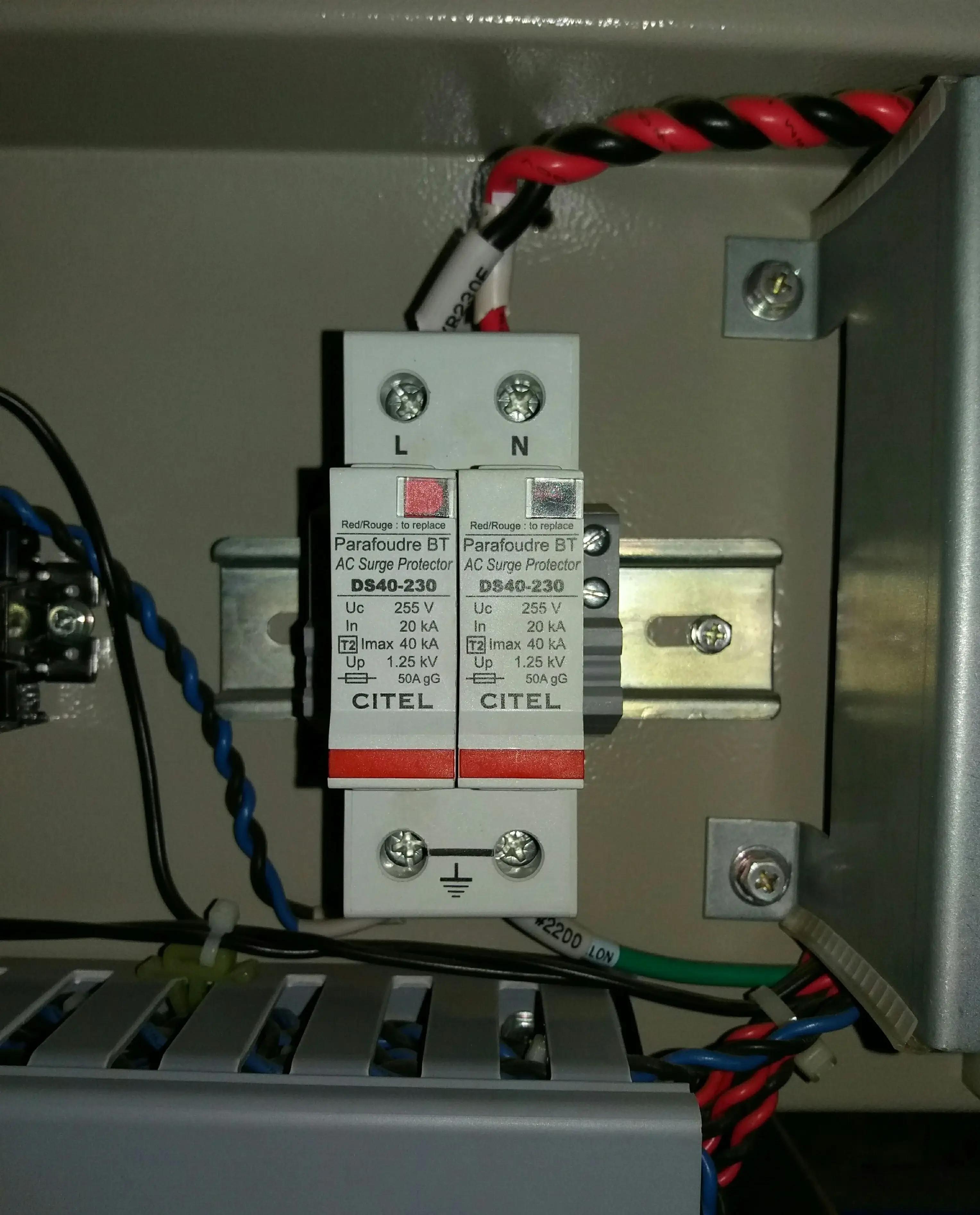 AC Surge Protector model DS40-230 installed on DIN rail in inverter AC panel, two modules for L and N conductors, left module status indicator window showing red meaning it has operated and needs replacement, right module shows grey operational status, twisted red and black cables entering from above