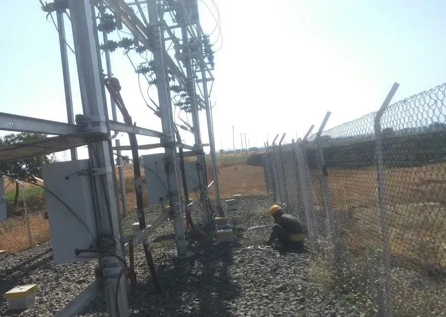 Solar plant technician in yellow helmet crouching at ground level inspecting surge arrester discharge counter or earthing junction boxes at base of HV substation structure, multiple white-painted square enclosures visible along the structure wall
