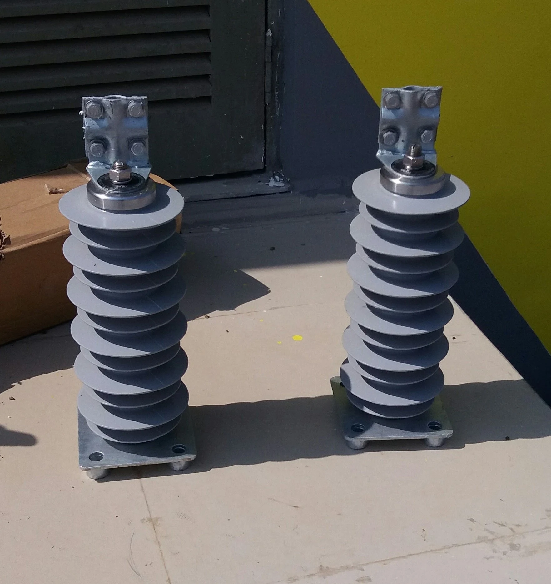 Two new grey polymer gapless lightning arresters with helical fin shed profile placed on tiled floor before installation at a solar plant HV substation, square base mounting plates visible