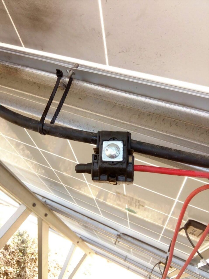 Insulation piercing connector joining black and red DC string cables on a solar panel frame rail beneath a ground-mounted PV array