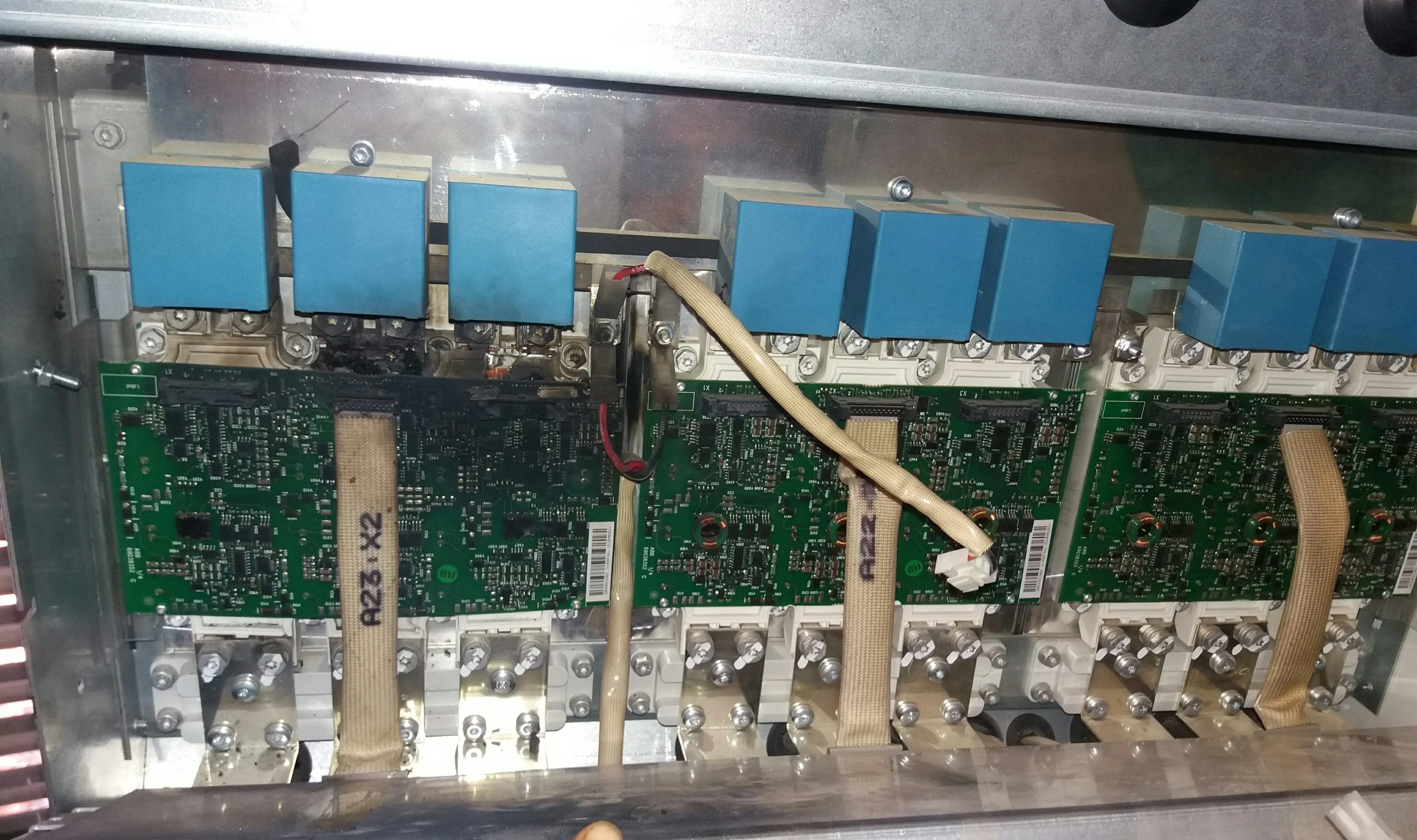 IGBT power module inside a central solar inverter power stage drawer showing severe burn and charring damage on the adjacent gate driver PCB, with blue snubber capacitors visible on top of undamaged neighbouring IGBT modules