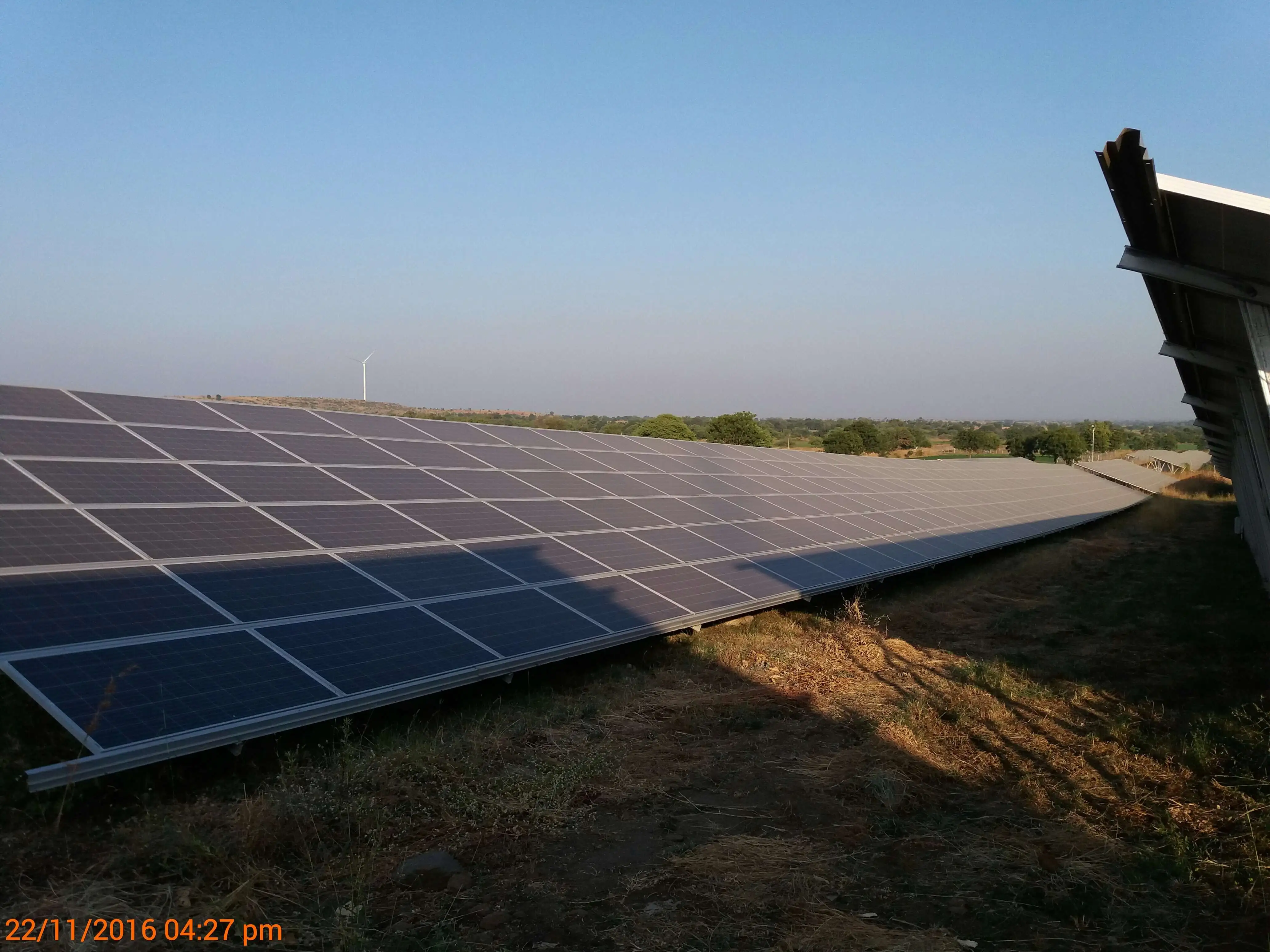 Wide view of a 15 MWp ground-mounted solar plant showing systematic inter-row shading across multiple array rows at 4:27 PM on 22 November 2016, with a wind turbine visible in the background