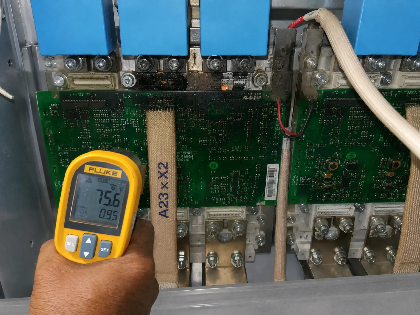 Fluke infrared thermometer held by a technician reading 75.6 degrees Celsius on the gate driver PCB surface of a central solar inverter power stage during post-failure inspection, emissivity set to 0.95, with visible charring on the PCB in the background